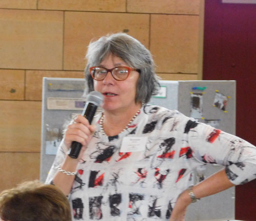 Woman with gray hair and red glasses stand with microphone.
