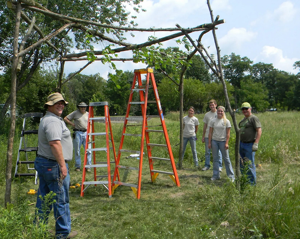 Six people standing around a structure made of branches