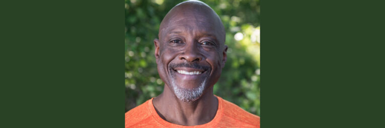 Headshot of Anthony Taylor in an orange shirt with leaves in the background