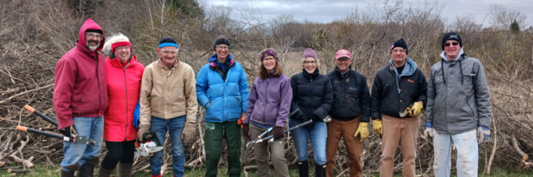 Nine people stand in front of a pile of cut branches, smiling.