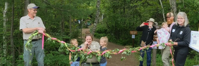 Members of the Friends of Tettegouche hold a ribbon intertwined with leaves while the assistant park manager prepares to cut it.