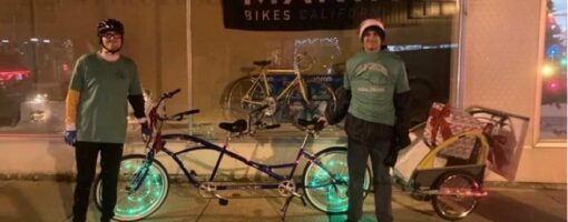 Two volunteers stand next to tandem bicycle with twinkle lights on wheels